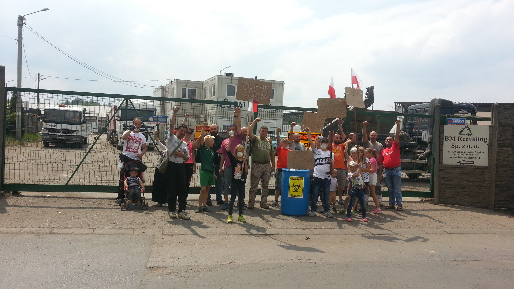 Grupa mieszkańców z dziećmi protestuje przed bramą zakładu BM Recykling Siemianowice, trzymając transparenty o zdrowiu i czystym powietrzu.